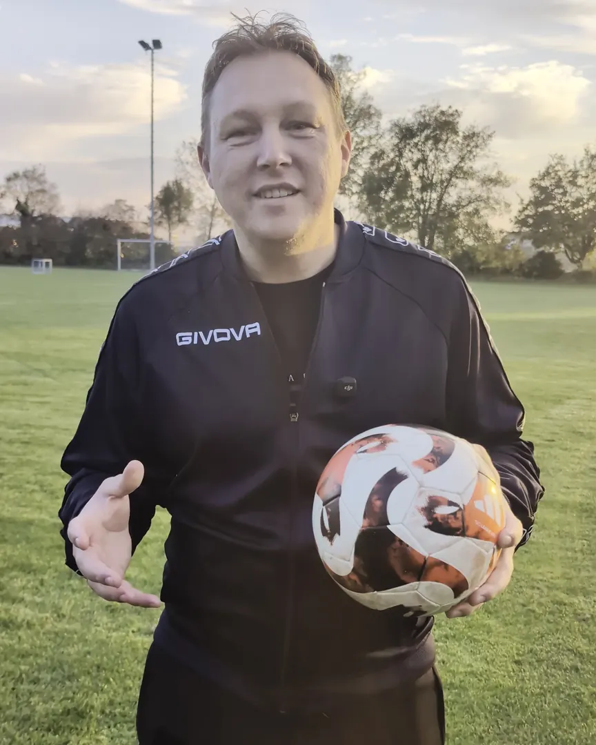 Stefan Eibl im schwarzen Trainingsanzug mit einem Ball unter dem Arm auf dem Sportplatz, im Hintergrund Abendhimmel und ein Flutlichtmast.