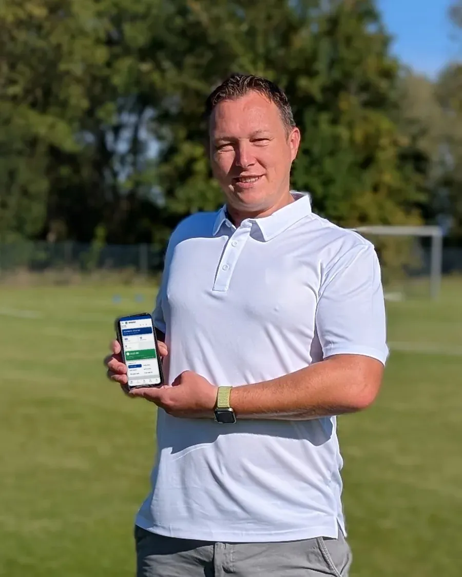 Stefan Eibl, Jugendleiter beim SV Pfatter, steht lächelnd auf dem Sportplatz in der Sonne und hält ein Handy mit Spielbereit in der Hand.